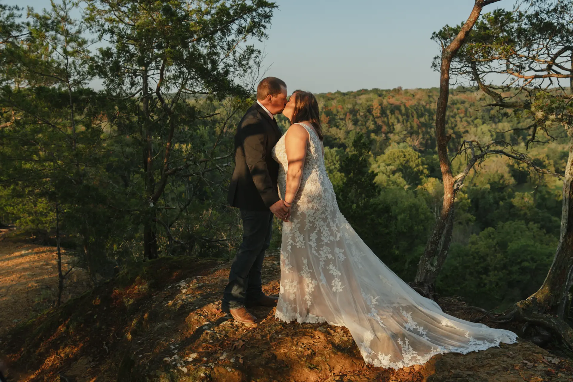 Sam and Justin kissing on a bluff overlook above the timber at sunset