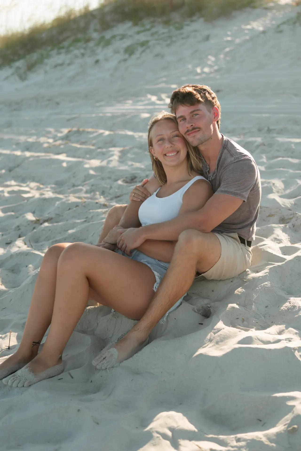 Bre and Luke sitting together in the sand at golden hour during their beach engagement session