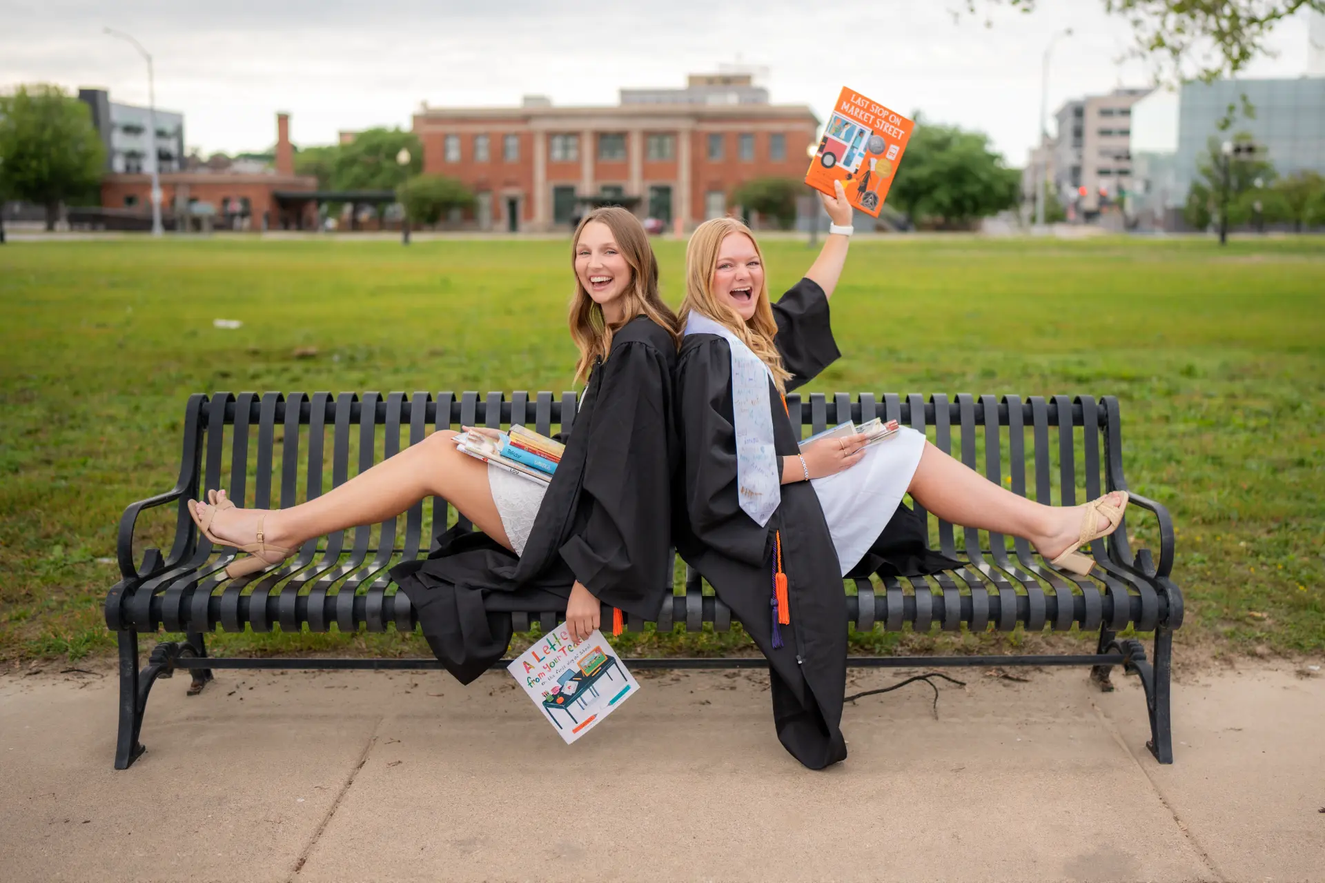 Two graduating seniors in caps and gowns laughing back to back on a park bench in downtown Davenport